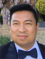 Photo of Henry Ocampo, MPH, wearing a dark jacket, light shirt, and a dark bow tie. In the background are a brick wall and purple flowers.