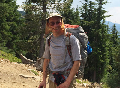 Dan Lowenstein hiking. He is smiling broadly and wearing a hat, sunglasses, large technical backpack, and gray t-shirt. Rocky terrain and evergreen trees are visible behind him, and mountain and sky in the distance.