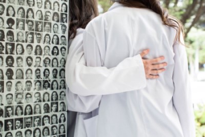 Two students wearing white coats, seen from behind with arms linked. A detail of the artwork, Columns, featuring portraits of individuals from UCSF’s history, is visible.